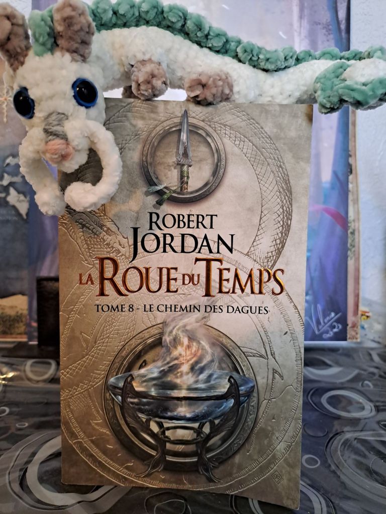 Photo montrant le livre La Roue du Temps, Le Chemin des dagues, de Robert Jordan. Il y a un genre de coupe sur la couverture. Il y a un dragon au crochet blanc et bleu (Haku) posé sur le livre. 