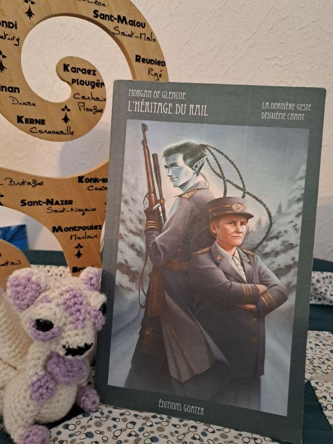 Photo montrant le livre 2 de la dernière geste (1 humain et un personnage bleuté sur la couverture, bordure de la couverture bleu-vert), à côté d'un renard au crochet.